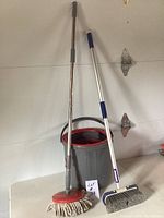 Photo of mop, bucket, and broom against a white wall, all standing upright.