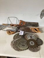 Multiple saw blades including a large handsaw with wooden handle, coping saw, and other handsaws arranged on a white surface with lot tag.