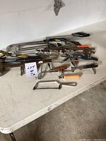 Photo showing multiple hand saws including hacksaws and coping saws, along with hammers and other hand tools laid out on a table.