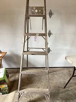 Front view of 6 foot aluminum step ladder showing multiple flat steps and folding metal frame, with garage door background.
