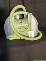 Front view of green Bissell Little Green upholstery cleaner showing hose wrapped on front with clear tanks on sides and carry handle on top.