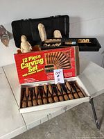 Overall view of 12 piece carving tool set with original box and unfinished carved wood pieces behind.