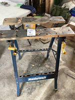 Front view of Mastercraft adjustable work bench showing wooden top with wear and blue metal frame with yellow adjustment knobs.