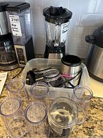 Shows Hamilton Beach blender with glass pitcher and black lid, Farberware stainless steel blender base, clear plastic blending containers, and blender accessories in plastic storage bin on kitchen counter.