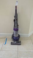 Front view of vacuum showing clear dust bin, purple ball base, and handle with controls on a tiled floor.
