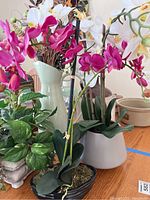 Photo showing pink orchid faux arrangement, leafy faux plant in stone pot, and tall vase with other faux flowers