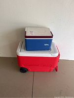 Photo showing a red Igloo cooler with wheels and a smaller blue cooler stacked on top, both with white lids and in need of cleaning.