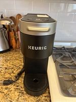 Front view of the black Keurig coffee maker placed on a kitchen counter near stove, showing power cord and drip tray.