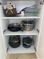 Three shelves within a white kitchen cabinet showing various pots, pans, and baking dishes arranged inside. The top shelf holds two rectangular glass baking dishes and a small basket alongside a red-handled pan. The middle shelf holds a frying pan, a stainless steel pan, and three metal pots with lids.