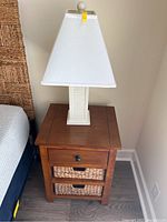 Wood night stand with two woven basket drawers and white lamp on top, full view showing overall condition and scratches on surface.