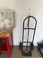 Milwaukee hand truck leaning against wall next to white box fan on red table.