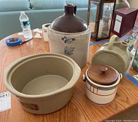 Photo of assorted pottery items on table including Western Stoneware crock, brown jug, round crock, small jar with wooden lid, and beige pitcher.