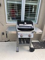 Front view of Weber 2-burner gas grill with side shelves and control knobs shown, outdoor setting on patio.