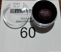 10 carat oval faceted ruby gemstone in a clear round container with black cushion, showing surface and internal inclusions.