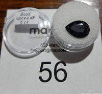 Blue sapphire gemstone in a round clear container with label 'Blue Sapphire 5ct'. Stone is pear shaped, dark blue with visible inclusions.