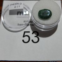 Oval green emerald gemstone placed on white foam inside clear container with label 'EMERALD 6 CT'.