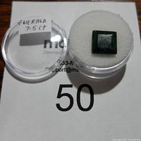 Top view of the emerald gemstone in a clear round container with a white foam pad and a label noting 7.5 carats.