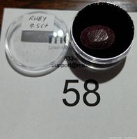Ruby gemstone in round clear plastic container showing label and gemstone inside black foam cushion.