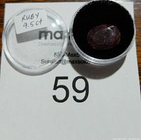 Clear plastic round container with black foam insert holding a 9.5 carat ruby gemstone with visible inclusions and the label 'Ruby 9.5 ct' on the lid.