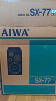 Photo of Aiwa speaker system box showing model SX-77 with product branding and speaker graphic illustration.
