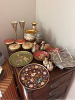 Photo showing enameled brass plates with intricate designs, brass vases, and duck figurines on wooden dresser.