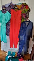 Photo shows five women's dresses and jumpsuits hanging on hangers, featuring teal, red, floral and polka dot designs.