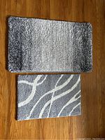 Two small area rugs laid flat on a wooden floor. One rug has a grey to white gradient pattern and measures 24 x 16 inches, the other has a grey background with white swirly lines and measures 20 x 32 inches.