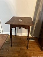 Full view of antique wooden side table showing square top, decorative apron, and four turned legs on hardwood floor against wall.