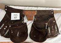 Full view of the brown leather tool belt laid flat, showing multiple large and small pockets riveted to the belt and overall wear including scuff marks and creasing.