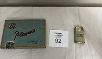 Photo of closed Player's cigarette tin with some writing on the lid and vintage Halifax butane lighter on the side.