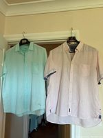 Two men's linen shirts hanging on hangers on a door, one light green Tommy Bahama and one pale apricot Marc O'Polo