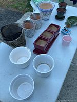 Multiple flower pots in various sizes and materials including ceramic, plastic, woven basket and novelty frog decorations shown on a table.