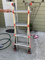 Folded aluminum ladder with orange highlights, wheels at the base, and metal rungs in front of a house exterior.