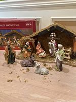 Photo of wooden moss manger and hand-painted ceramic nativity figures including Mary, Joseph, wise men, a woman, and animals donkey, cow, sheep. Figures arranged in front of manger for display.