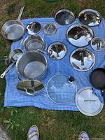 Overview of pots, pans, and numerous lids arranged on a blue checkered cloth on grass, showing a variety of sizes and reflective metal finishes.