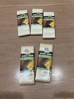 Five unopened packages of satin blanket binding, three white and two oyster, displayed on wooden surface.