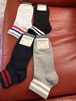 Four pairs of Kikiyasocks men's ankle socks displayed on a red surface, showing white, black, navy blue, and grey colors with striped cuffs, each pair attached to its packaging tag.