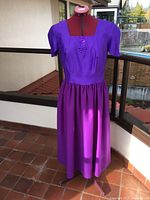 Short-sleeved purple dress shown on dress form indoors on tiled floor with balcony background.