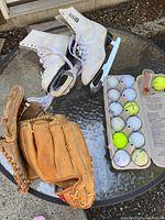 Photo shows the vintage CCM figure skates, two leather baseball gloves, and assorted golf balls with packaging on a glass table.