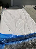 Mattress wrapped in clear plastic with blue bottom layer, folded on driveway.