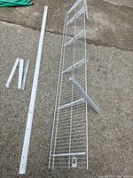 Photo of white metal wire shelf placed on ground showing horizontal grid bars and a wall bracket attached underneath.