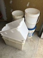 3 white plastic storage bins stacked with several white plastic buckets nearby on a concrete floor in a garage or storage space