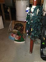 Photo of assorted Christmas decorations including string lights, wreath, gift bags, stockings, and tree skirt placed on basement floor near water heater and pipe.