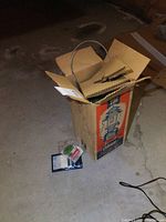 Cardboard box upright on floor showing Coleman 236 box graphic and spare wick packets on the ground beside it.