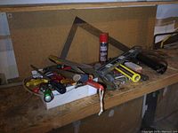 Wide view of mixed hand tools on wooden workbench with pegboard background, including screwdrivers, hammers, scraper, and spray can.