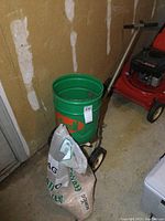 Photo of green So-Green branded lawn spreader with metal frame and wheels next to a garage wall and placed near a red lawn mower.