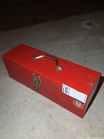 Closed red metal toolbox on concrete floor, showing rectangular shape with handle and latch.
