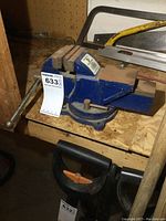 Blue swivel base bench vise on a wooden surface with dust and signs of wear.