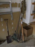 Wide view showing multiple yard tools grouped against wall including shovel, rake, and rope
