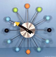 Full front view of the colorful George Nelson atomic wall clock showing all twelve sphere markers and black hands with yellow arrow.